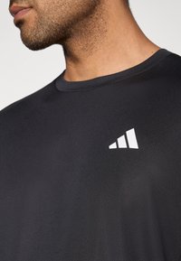 Svart atletisk t-skjorte med rund hals; har et teksturert stoff og en hvit logo med tre striper på venstre side nær skulderen.