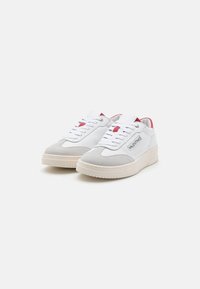 Sneaker bianche e grigie con punta in suede texturizzato, tomaia in pelle liscia, dettagli rossi e suola in gomma con piccole indentazioni.