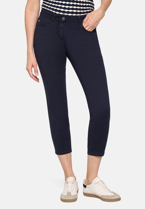 Kvinde iført marineblå, korte, slanke bukser, stribet top og hvide sneakers, stående med den ene hånd i lommen foran en hvid baggrund.