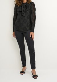 Kvinna iklädd svart spetsblus med lång ärm och rosett, slimmade svarta byxor och svarta klacksandaler med flätade remmar, stående mot en enfärgad bakgrund.