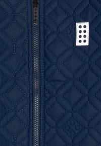 Marineblauer, gesteppten Jacke mit zentralem Reißverschluss, Rauten-Stitching und einem weißen Etikett mit schwarzen Punkten auf der linken Seite.