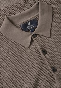 Camisa polo marrón texturizada con un patrón ondulado, con tapeta de botones y cuello. Cuenta con dos botones negros y una etiqueta de tela que muestra el nombre de la marca.