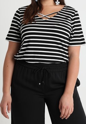 Kobieta w czarno-białym poziomo paskowanym topie z krótkim rękawem i dekoltem w kształcie krzyża, w czarnych spodniach z troczkami, z widocznymi białymi kolczykami w kształcie kwiatu.