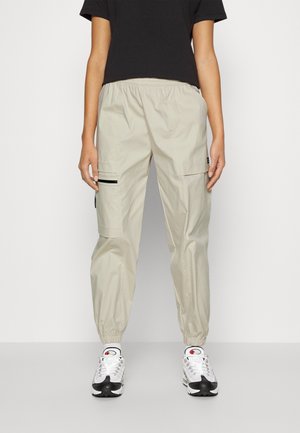 Person trägt beige Cargo-Hose mit elastischen Bündchen, schwarzes kurzes T-Shirt und weiße Sportschuhe, steht vor einem einfarbigen Hintergrund.