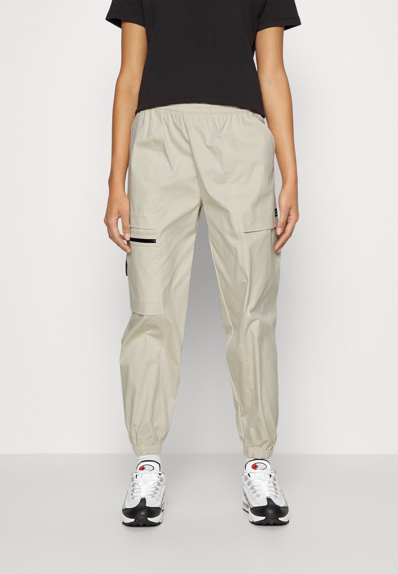 Personne portant un pantalon cargo beige avec des chevilles élastiques, un t-shirt court noir et des chaussures de sport blanches, debout devant un fond uni.