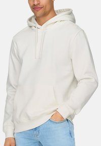 Haut à capuche crème avec une capuche à cordons, poche kangourou, poignets et ourlet côtelés, fabriqué en tissu doux et texturé, associé à un jean bleu clair.