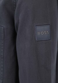 Marineblauwe jas van textuurstof, met op de mouw een vierkante patch met de merknaam "BOSS" in een contrasterende kleur.