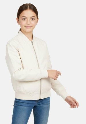 Hellbeige Bomberjacke mit einem Frontreißverschluss, gerippten Bündchen und Saum; kombiniert mit blauen Jeans. Das Model posiert mit einer Handgeste.