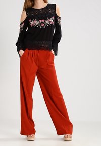 Svart kallskulderblus med blommig broderi; röda vida byxor; texturerad tyg; spetsdetalj i fållen. Sandaler fulländar outfiten.