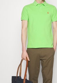 Polo verde con colletto e maniche corte. Pantaloni marroni e una borsa a tracolla blu con spalline beige. Design semplice con un piccolo logo.