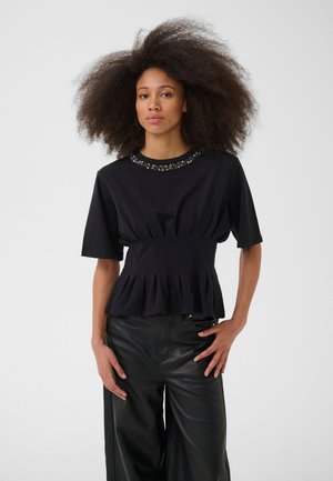 Jeune femme aux grands cheveux bouclés portant un haut noir à manches courtes avec un col orné de perles et un pantalon large en cuir noir.