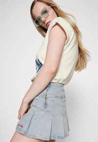 Saia mini de denim com pregas, na cor azul clara, apresentando um fecho zíper nas costas. Combinada com uma camisa de mangas curtas de cor creme e óculos de sol verdes.
