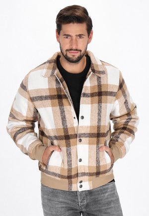 Man wearing a beige and white plaid jacket with front pockets, standing with hands in pockets and looking forward against a plain background.