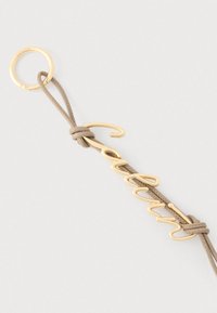 Μπεζ δερμάτινο μπρελόκ με χρυσό μεταλλικό κρίκο και χρυσό λογότυπο "calvin" σε καλλιγραφικό στυλ, προσαρτημένο με κόμπους και στις δύο άκρες του λουριού.