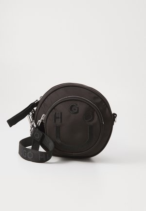 Petit sac bandoulière rond noir avec poche zippée à l'avant, lettres brodées et bandoulière réglable avec fermoirs métalliques.