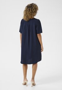 Tummansininen polvipituinen mekko, jossa on lyhyet hihat, teksturoitu kangas, pleatattu selkäosa, yhdistettynä vaaleansinisiin korkokenkiin.