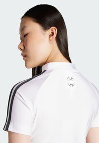 Biela športová košeľa s čiernymi pruhmi na pleciach. Má vysoký golier a okrúhly logo na zadnej strane blízko výstrihu.