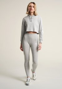 Ljusgrå cropped hoodie med snörning och logotyp, kombinerad med matchande högmidjade leggings. Modellen bär grå och vita sneakers.