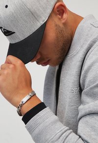 Grijze zip-up sweatshirt met zwarte accenten, grijze baseballcap en zilveren armband. Close-up van een hand op de kin, die de accessoires toont.