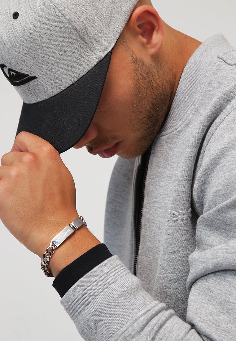 Grijze zip-up sweatshirt met zwarte accenten, grijze baseballcap en zilveren armband. Close-up van een hand op de kin, die de accessoires toont.