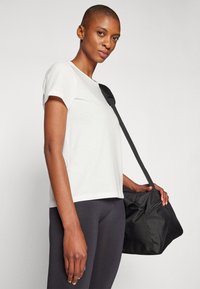 White short-sleeve t-shirt in soft fabric, paired with black leggings. A black duffle bag with a shoulder strap is held in one hand.