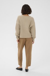 Pull en maille beige à la coupe ample, avec un col et des poignets côtelés. Associé à un pantalon brun ample et des chaussures noires.