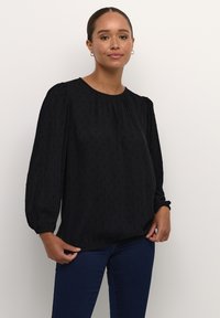 Blouse noire à manches bouffantes en tissu texturé avec un motif subtil. Elle présente un col rond et des poignets élastiques, associée à un jean foncé.