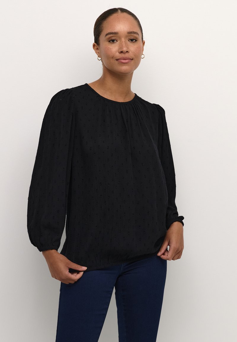 Blouse noire à manches bouffantes en tissu texturé avec un motif subtil. Elle présente un col rond et des poignets élastiques, associée à un jean foncé.