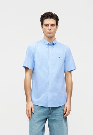 Νέος άνδρας με καστανά μαλλιά, που φοράει πουκάμισο κοντομάνικο ανοιχτού μπλε και λευκών ριγών και ανοιχτό μπλε τζιν παντελόνι, κοιτάζει μπροστά με ουδέτερη έκφραση.