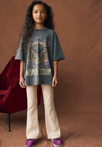 Graues, übergroßes T-Shirt mit Schmetterlings- und Blumenmotiven, Text "WORLD TOUR", kombiniert mit hellbeigen Schlaghosen und lila Sneakers.