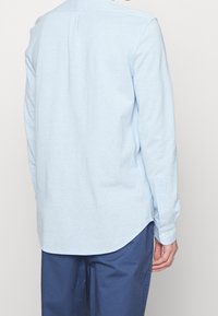 Světle modrá, dlouhá košile s zaobleným lemem a knoflíkovými manžetami. Vyrobena z měkké tkaniny, s jednoduchým designem a detailem s plisem na zadní straně.