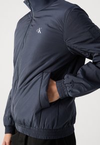 Granatowa lekka kurtka z wysokim kołnierzem, zamkiem błyskawicznym, elastycznymi mankietami, bocznymi kieszeniami oraz logo na piersi i rękawie.