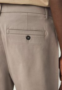Beige Stoffhose mit einer Gesäßtasche, die mit einem schwarzen Knopf gesichert ist, und einer glatten Textur sowie tailliertem Design.