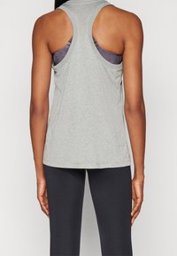 Kobieta w szarej, bezrękawnikowej koszulce do ćwiczeń na purpurowym biustonoszu sportowym i czarnych legginsach, pokazana z tyłu na jednolitym tle.