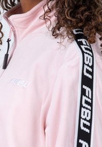 Chaqueta de forro polar rosa con cuello alto, rayas laterales en blanco y negro con el logo "FUBU" y el logo "FUBU" bordado en el pecho.