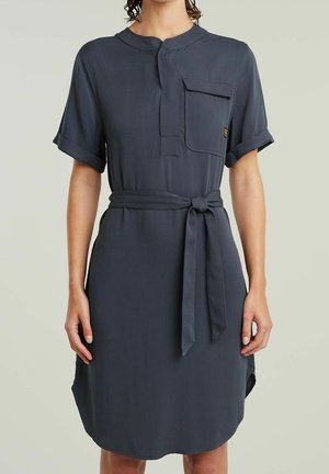 Femme portant une robe ceinturée gris foncé à manches courtes avec une poche sur la poitrine et des poignets retroussés, debout devant un fond clair uni.