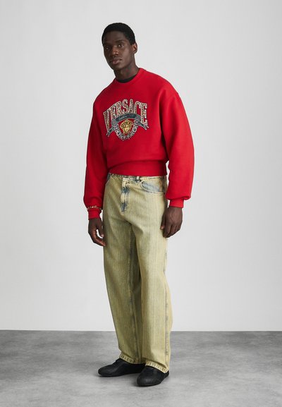 Punane sü sweatshirt, millel on graafiline kujundus koos lõviga, kombineeritud heledate kotikeste denimpükstega ja mustade tossudega.