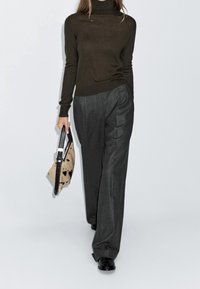 Femme marchant, portant un col roulé marron foncé et un pantalon large gris, tenant un sac à main beige à pois noirs avec une bandoulière noire.