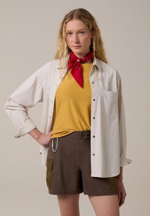 Mujer con cabello largo y rubio que lleva una bufanda roja en el cuello, camisa amarilla, camisa beige abotonada y pantalones cortos marrones con un mosquetón.