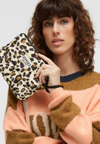 Quilted clutch in cream with brown and black leopard print. Features a logo patch and a wrist strap. Model wearing a multicolor sweater.