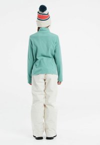 Pullover in pile turchese con colletto alto, abbinato a pantaloni da neve bianchi larghi. Un berretto a maglia a righe con pompon blu adornano la testa.