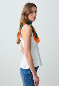 Jongemeisje met lang haar, gekleed in een witte mouwloze top, blauwe jeans en een oranje en groene sjaal om haar hals, kijkend opzij.