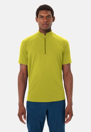 Man staat rechtop met het gezicht naar voren, draagt een limegroen shirt met korte mouwen en ritssluiting, en blauwe broek, tegen een effen achtergrond.