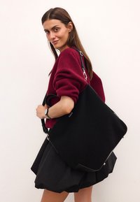 Czarna torba tote z płótna z srebrnym wykończeniem, w strukturze utwardzonej z bocznymi uchwytami, noszona przez modelkę w burgundowym swetrze.