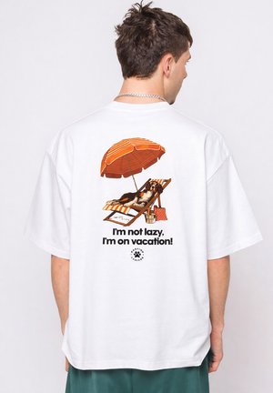 Homme portant une chemise blanche avec une illustration de chien se détendant sur une chaise rayée sous un parasol orange et le texte "Je ne suis pas paresseux, je suis en vacances !"