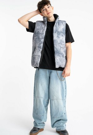 Młoda osoba stojąca z jedną ręką na głowie, ubrana w szarą pikowaną kamizelkę w wzory, czarną koszulkę, szerokie jasnoniebieskie dżinsy i buty sportowe.