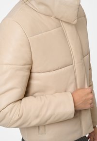 Beige läder puffjacka med hög krage, quiltat mönster och sidofickor. Slät textur; skräddarsydd passform.