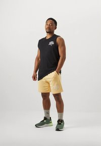 Camiseta preta sem mangas, calções amarelos com logótipo branco, meias cinzentas, sapatos de desporto verdes e pretos. Pose em pé contra um fundo branco.