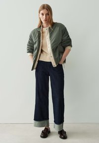 Olivgrüne Bomberjacke über beigem Strickpullover, kombiniert mit weit geschnittenen, dunkelblauen Jeans mit hellblauen Säumen und braunen Slippern.