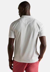 Balts polo krekls ar ribinātu apkakli un īsām piedurknēm, kam ir smalka tekstūra un taisns apakšmalas garums. Valkāts ar rozā šortiem.